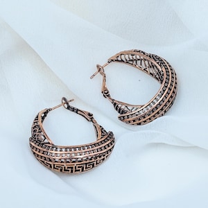 May include: A pair of copper-toned hoop earrings featuring an intricate openwork design with a repeating Greek key pattern along the bottom edge and a textured, dotted grid motif across the upper curved surface, secured with a hinged post closure.