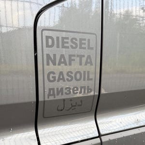 Könnte beinhalten: Eine graue Fahrzeugtür mit einem rechteckigen Aufkleber mit der Aufschrift "DIESEL, NAFTA, GASOIL" auf Englisch, gefolgt von Text in kyrillischer und arabischer Schrift. Die Tür hat einen kleinen kreisförmigen Sensor.