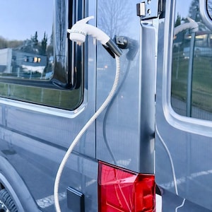 Könnte beinhalten: Außenansicht eines grauen Transporters mit einem weißen Duschkopf und Schlauch, die an der Seite befestigt sind. Der Transporter hat getönte Scheiben und ein rotes Rücklicht. Der Duschkopf ist in der Nähe der Tür montiert.