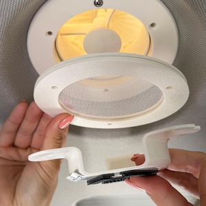 Op de afbeelding: Witte plafondventilatie met een cirkelvormig ontwerp. De ventilatie heeft een gaasscherm en wordt door een persoon vastgehouden. Een aparte witte beugel met een zwarte strip is ook zichtbaar.