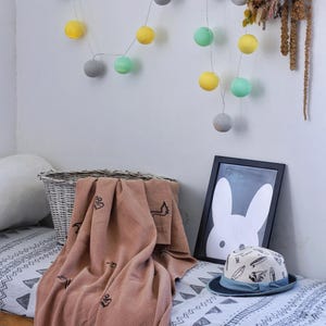 May include: A cozy scene featuring a light brown blanket with black embroidered designs, a framed bunny print, and a patterned hat. A string of colorful, round lights hangs on the wall, adding a playful touch to the room's decor.
