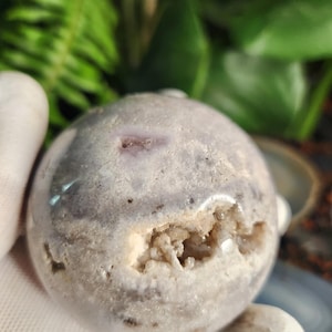 May include: A spherical, light gray crystal geode with a cavity revealing a cluster of small, sparkling crystals. The geode has a smooth, polished surface with subtle variations in color and texture. The background is blurred with green foliage.
