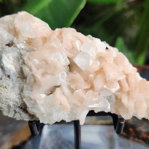 Op de afbeelding: Een close-up van een ruw mineraal specimen. De steen heeft lichtroze en heldere kristalformaties die zijn gegroepeerd op een grijze matrix. De kristallen hebben een sprankelende, doorschijnende kwaliteit en de algehele textuur is ruw en ongelijkmatig.