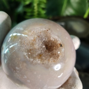 May include: A polished, spherical geode with a light purple and white exterior. The geode has a cavity filled with sparkling crystals and a rough, textured interior. The sphere is held in a gloved hand, with green foliage in the background.