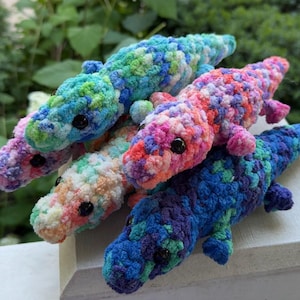 May include: Five crocheted alligator plush toys in various colours, including blue, pink, and green. Each toy has black button eyes. The toys are arranged on a white surface, with foliage visible in the background. The toys are handmade.