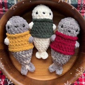 Puede incluir: Tres peluches de foca hechos a mano con ganchillo en un cuenco de madera. Las focas tienen cuerpos grises y visten jerséis amarillos, verdes y rojos. Los peluches tienen ojos negros y vientres blancos. El cuenco está sobre una superficie a cuadros.