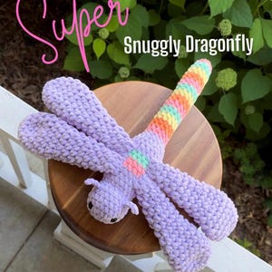 May include: A crochet dragonfly plush toy featuring a lavender body and four large lavender wings. The tail is striped with rainbow colors including pink, orange, yellow, green, and blue. The text Super Snuggly Dragonfly is printed in pink and white.