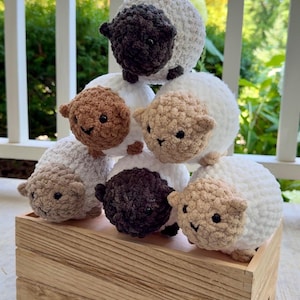 May include: A collection of handmade crochet sheep in various colors, including white, brown, and gray, are arranged in a wooden box. Each sheep has a smiling face and small ears, creating a whimsical and charming display.