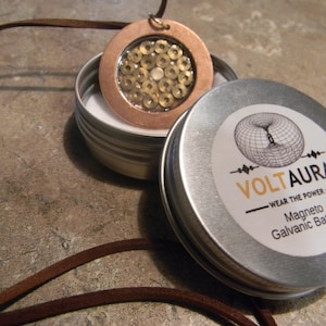 May include: A copper pendant with a clear centre filled with small, gold-coloured beads, displayed with a metal tin. The tin lid features the text "VOLTAURA" and "Magneto Galvanic Battery". A brown leather cord is attached to the pendant.