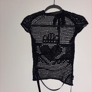 May include: Black crochet top with short sleeves, hanging on a hanger. The open weave design reveals the background. The top has a round neckline and a ribbon detail at the back. The garment is made of a dark, possibly cotton, yarn.