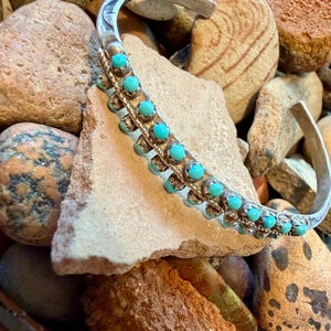 May include: A silver cuff bracelet featuring two parallel rows of small, round, bright turquoise stones set in scalloped metal bezels. The band has a twisted wire design between the rows of stones and etched detailing on the ends of the cuff.