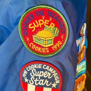 Puede incluir: Chaqueta azul con m&uacute;ltiples parches. Los parches incluyen texto: "COOKIE FUN FAIR 89 Super Gruntastic", "DISCOVER THE TREASURES SUPER COOKIES 1990", y "COOKIE CAMPAIGN Super Star".