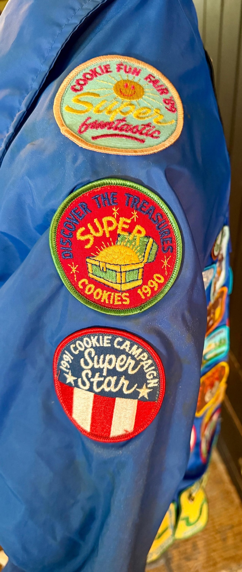 Puede incluir: Chaqueta azul con m&uacute;ltiples parches. Los parches incluyen texto: "COOKIE FUN FAIR 89 Super Gruntastic", "DISCOVER THE TREASURES SUPER COOKIES 1990", y "COOKIE CAMPAIGN Super Star".