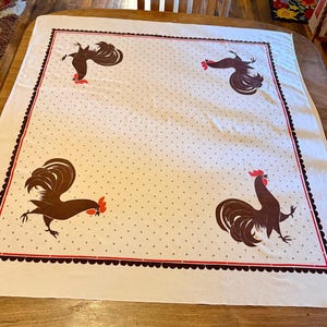 May include: A square white tablecloth featuring a repeating pattern of small gray polka dots. The center area is framed by a dark brown scalloped border and a thin red line. Four brown roosters with red combs are positioned in the corners.