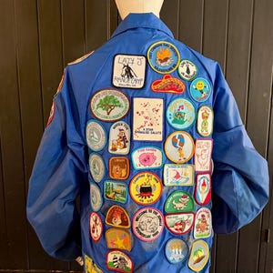 Puede incluir: Una chaqueta azul cubierta de parches coloridos. Los parches presentan varios dise&ntilde;os, incluyendo un cerdo, un &aacute;rbol y texto que dice "Lazy J Ranch Camp" y "Girl Scouts". La chaqueta tiene cuello y mangas largas.