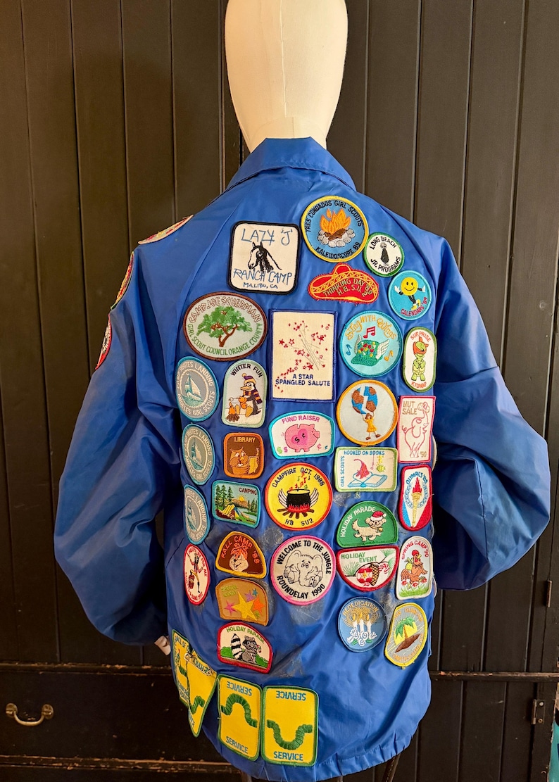 Puede incluir: Una chaqueta azul cubierta de parches coloridos. Los parches presentan varios dise&ntilde;os, incluyendo un cerdo, un &aacute;rbol y texto que dice "Lazy J Ranch Camp" y "Girl Scouts". La chaqueta tiene cuello y mangas largas.