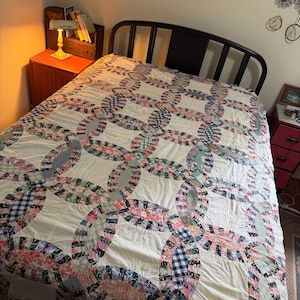 May include: A double wedding ring pattern quilt featuring a white background with interlocking rings made of various multicolored fabric scraps, including floral, plaid, and geometric prints, covering a bed with a black metal frame.