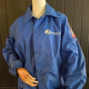 Puede incluir: Chaqueta azul de nailon con cierre de botones y cuello. La chaqueta presenta el logotipo y el texto de las Girl Scouts en el pecho y un parche en la manga. La chaqueta se exhibe en un maniqu&iacute;.