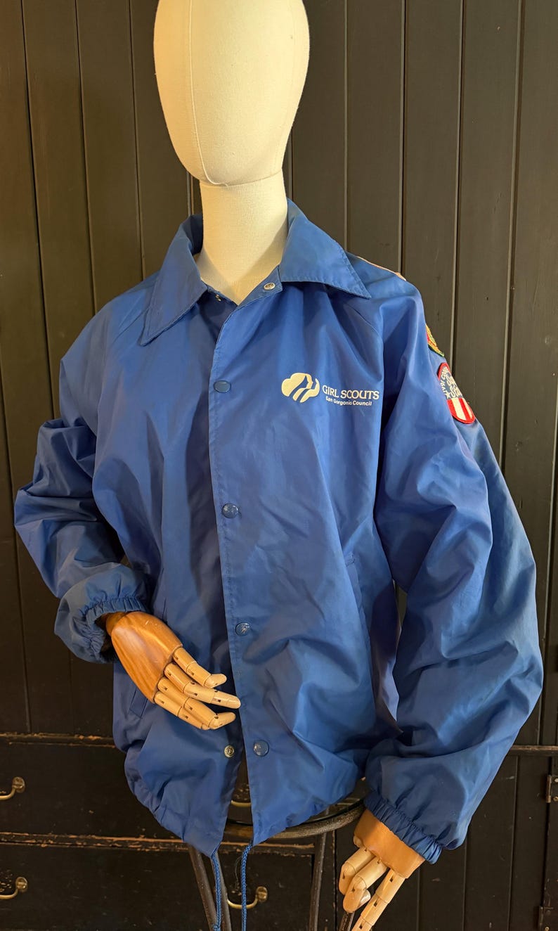Puede incluir: Chaqueta azul de nailon con cierre de botones y cuello. La chaqueta presenta el logotipo y el texto de las Girl Scouts en el pecho y un parche en la manga. La chaqueta se exhibe en un maniqu&iacute;.