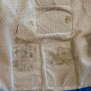 Puede incluir: Primer plano de una tela acolchada con una superficie texturizada de color gris claro. La tela tiene varios parches rectangulares cosidos. Los parches tienen una decoloraci&oacute;n m&aacute;s oscura. Un borde azul es visible en la parte inferior.