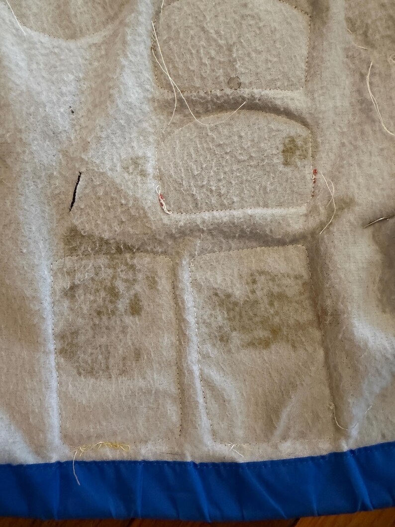 Puede incluir: Primer plano de una tela acolchada con una superficie texturizada de color gris claro. La tela tiene varios parches rectangulares cosidos. Los parches tienen una decoloraci&oacute;n m&aacute;s oscura. Un borde azul es visible en la parte inferior.