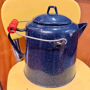 May include: A dark blue enamel coffee pot with a white speckled pattern, featuring a matching lid, a metal handle with a red wooden grip, and a side spout. The cylindrical vessel has a sturdy construction and a classic design for brewing.
