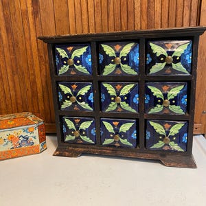 May include: A dark wooden cabinet with nine drawers, each adorned with a blue tile featuring a floral design in green, gold, and blue. The cabinet has a distressed finish, and a decorative tin is visible to the left.