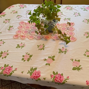 May include: A white tablecloth featuring a repeating pattern of pink roses with green leaves, topped with a rectangular pink and white crocheted doily and a potted green oxalis plant with triangular leaves in a gray square container.