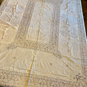 May include: A large, off-white tablecloth with intricate lace detailing. The design features a central rectangular panel surrounded by a border of floral and paisley patterns. Smaller rectangular panels are placed around the center. The edges are finished with a delicate lace trim.