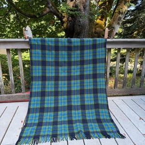 May include: A rectangular wool blanket featuring a plaid pattern in shades of dark navy blue, bright blue, and olive green. The blanket has a textured weave and is finished with a fringe along the bottom edge, displaying a consistent grid design.