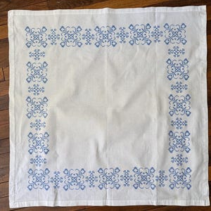May include: A square white linen tablecloth featuring a blue cross-stitch border pattern. The design consists of repeating geometric floral motifs arranged in a continuous frame around the perimeter, with a plain white center and hemmed edges.