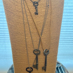 May include: A display of antique key necklaces on a mannequin. The necklaces feature various key designs on ball chains and leather cords. Several additional keys are arranged at the base, showcasing a vintage aesthetic. The mannequin is set on a rustic wooden surface.