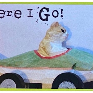 Puede incluir: Una caprichosa figura de gato en un coche con forma de pez con el texto "Here I Go!". El gato es marr&oacute;n claro y blanco, y el coche es verde, blanco y negro. El coche pez tiene ruedas negras y un ribete rojo. El fondo es azul claro.