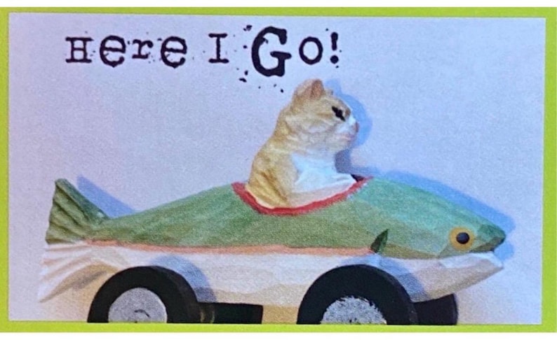 Puede incluir: Una caprichosa figura de gato en un coche con forma de pez con el texto "Here I Go!". El gato es marr&oacute;n claro y blanco, y el coche es verde, blanco y negro. El coche pez tiene ruedas negras y un ribete rojo. El fondo es azul claro.