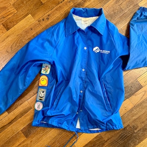 Puede incluir: Chaqueta azul brillante de las Girl Scouts con cierre de botones, cuello y mangas largas. La chaqueta presenta el logotipo de las Girl Scouts y varios parches coloridos en la manga izquierda. La chaqueta es de material ligero.