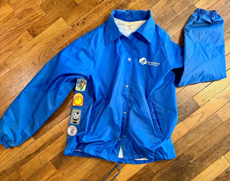 Puede incluir: Chaqueta azul brillante de las Girl Scouts con cierre de botones, cuello y mangas largas. La chaqueta presenta el logotipo de las Girl Scouts y varios parches coloridos en la manga izquierda. La chaqueta es de material ligero.