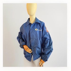 Puede incluir: Chaqueta azul de nailon con el logotipo de las Girl Scouts en el pecho y parches en la manga. La chaqueta tiene cierre de botones, cuello y pu&ntilde;os el&aacute;sticos. La chaqueta se exhibe en un maniqu&iacute;.
