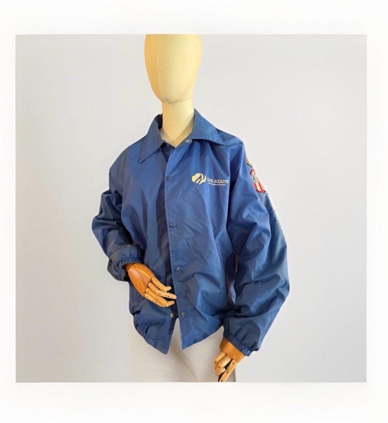 Puede incluir: Chaqueta azul de nailon con el logotipo de las Girl Scouts en el pecho y parches en la manga. La chaqueta tiene cierre de botones, cuello y pu&ntilde;os el&aacute;sticos. La chaqueta se exhibe en un maniqu&iacute;.