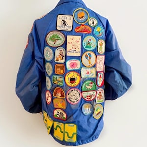 Puede incluir: Chaqueta azul de nailon cubierta de parches bordados coloridos. Los parches presentan varios dise&ntilde;os, incluyendo logotipos de campamentos, personajes de dibujos animados y texto. La chaqueta tiene un estilo bomber cl&aacute;sico.