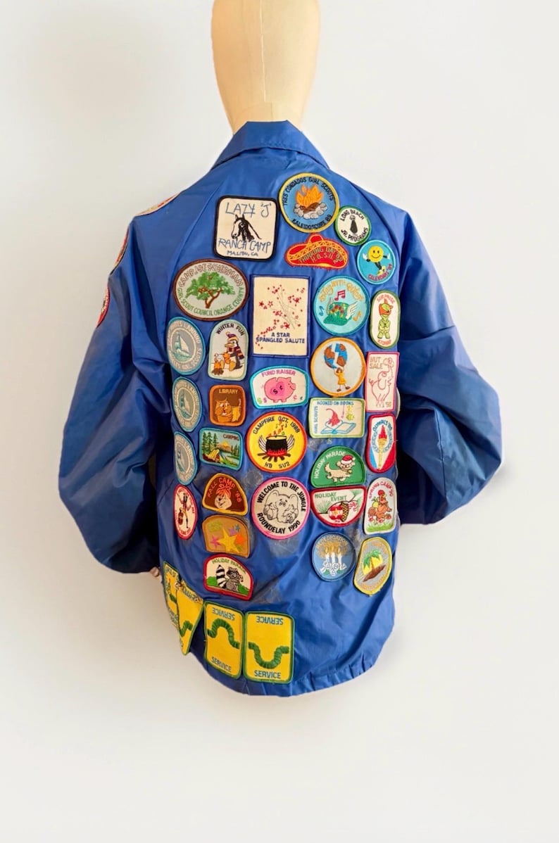 Puede incluir: Chaqueta azul de nailon cubierta de parches bordados coloridos. Los parches presentan varios dise&ntilde;os, incluyendo logotipos de campamentos, personajes de dibujos animados y texto. La chaqueta tiene un estilo bomber cl&aacute;sico.