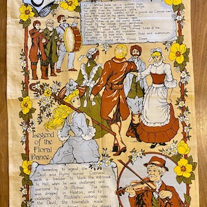 May include: A beige cotton tea towel featuring a colorful illustration of the Cornish Floral Dance. The design includes figures in period clothing, musicians, and floral accents. Text details the dance's legend and history.