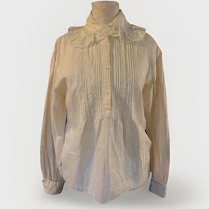 May include: Cream-colored long-sleeve blouse with a high collar and a lace-trimmed ruffle. The front features vertical pleats and small, round buttons. The sleeves have buttoned cuffs. The blouse is displayed on a mannequin.