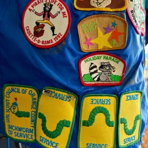 Puede incluir: Una colecci&oacute;n de parches bordados coloridos sobre un fondo azul. Los parches incluyen un pirata, un mapache y un gusano. El texto incluye "A Pirate's Life For Me" y "Service".
