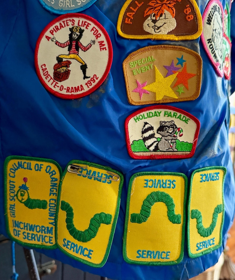 Puede incluir: Una colecci&oacute;n de parches bordados coloridos sobre un fondo azul. Los parches incluyen un pirata, un mapache y un gusano. El texto incluye "A Pirate's Life For Me" y "Service".