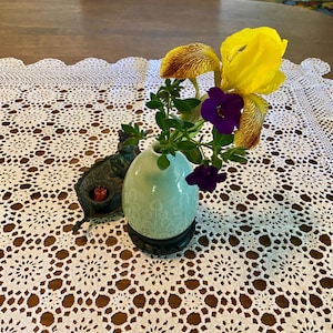 May include: A small, light blue vase with a floral design holds a yellow iris and purple flowers. The vase sits on a black stand, and is placed on a white lace tablecloth. A decorative metal cat figurine is to the left of the vase.