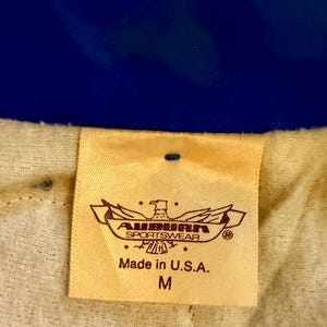 Puede incluir: Primer plano de una etiqueta beige con el texto "AUBURN SPORTSWEAR" y "Made in U.S.A.". La etiqueta tambi&eacute;n presenta un emblema de &aacute;guila y la talla "M". El fondo es una tela azul.