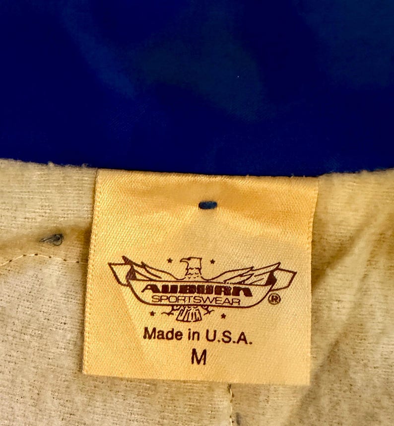 Puede incluir: Primer plano de una etiqueta beige con el texto "AUBURN SPORTSWEAR" y "Made in U.S.A.". La etiqueta tambi&eacute;n presenta un emblema de &aacute;guila y la talla "M". El fondo es una tela azul.