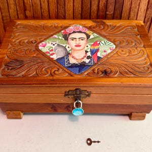 May include: A rectangular wooden box with carved scrollwork patterns on the lid and a central diamond-shaped inlay featuring a portrait of a woman with a floral crown, a lemur, and a parrot. The box has a metal latch with a blue padlock and a small key.