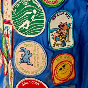 Puede incluir: Una colecci&oacute;n de parches coloridos de Girl Scout sobre un fondo azul. Los parches presentan varios dise&ntilde;os, incluyendo una figura corriendo, un oso en la playa, y textos como "Holiday Event" y "Fun Day".