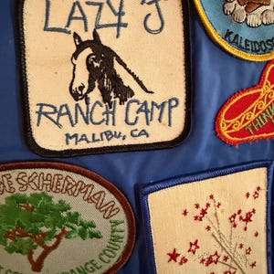 Puede incluir: Colecci&oacute;n de parches bordados sobre fondo azul. Uno de los parches dice "LAZY J RANCH CAMP MALIBU, CA" con una ilustraci&oacute;n de un caballo. Otros parches presentan texto e ilustraciones, incluyendo una fogata, estrellas y un &aacute;rbol.